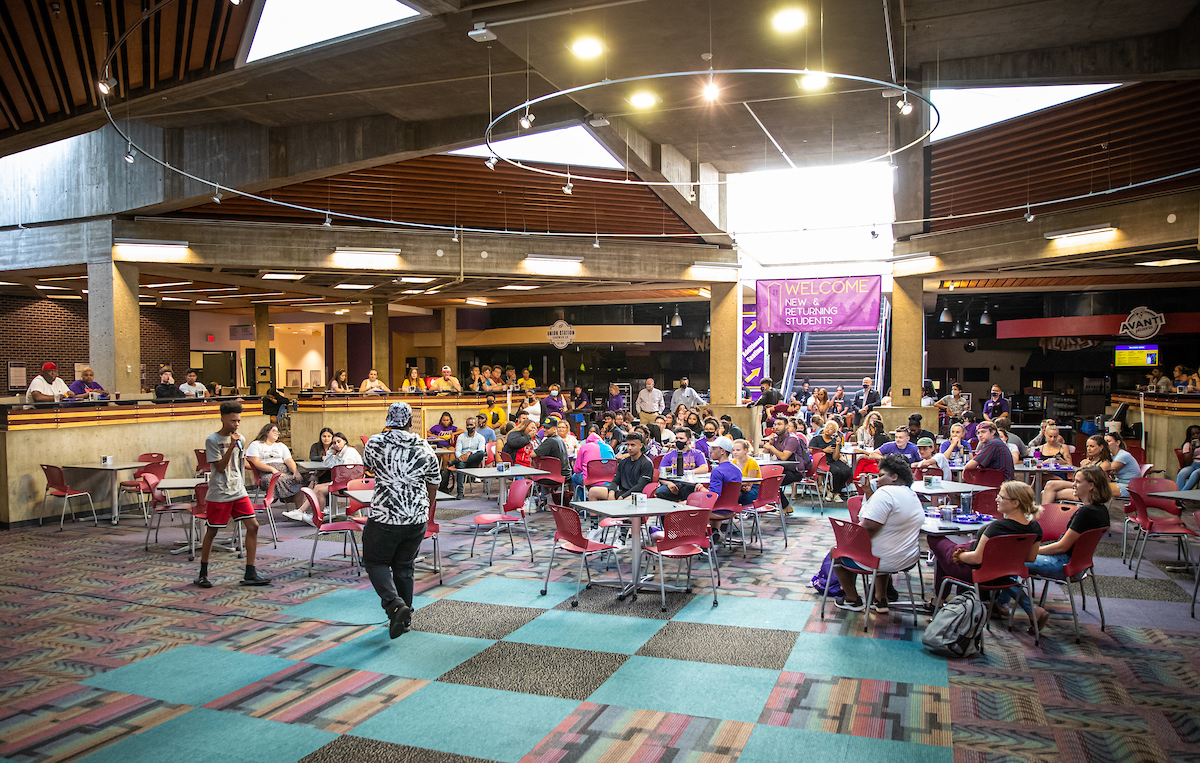 Students sitting at tables in the union listening to a performer. Sign in background reads welcome new and returning students.